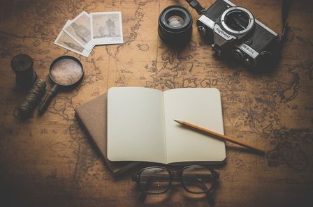 Carnet aux pages blanches avec un crayon de bois, entouré d'une loupe, d'un appareil photo ancien et de divers objet, posés sur une carte ancienne du monde