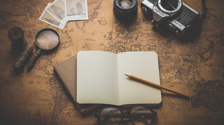 Carnet aux pages blanches avec un crayon de bois, entouré d'une loupe, d'un appareil photo ancien et de divers objet, posés sur une carte ancienne du monde