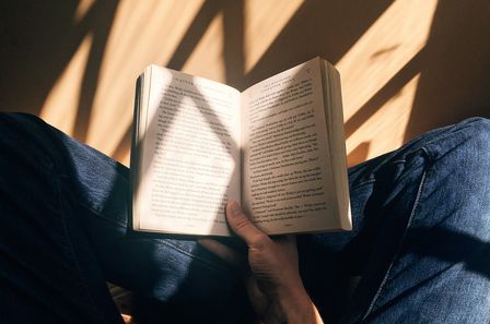 Livre ouvert sur les genou d'un homme assis en tailleur, portant un jean, à la lumière d'une fenêtre