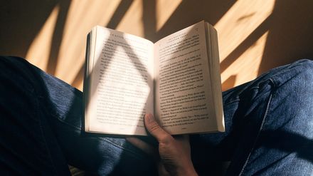 Livre ouvert sur les genou d'un homme assis en tailleur, portant un jean, à la lumière d'une fenêtre