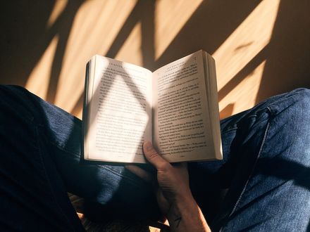 Livre ouvert sur les genou d'un homme assis en tailleur, portant un jean, à la lumière d'une fenêtre