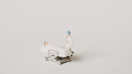 2 petits jouets représentant un chirurgien et son patient alité, sur fond blanc