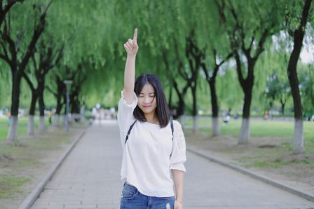 Jeune femme les yeux fermés et souriante, pointant le doigt vers le ciel sur fond de parc