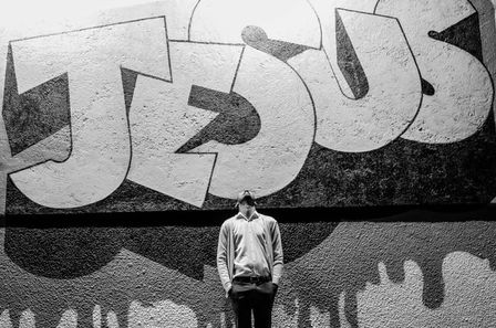Photo en noir & blanc d'un homme le visage au ciel devant un mur où se trouve un immense tag "Jésus"