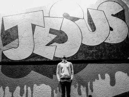 Photo en noir & blanc d'un homme le visage au ciel devant un mur où se trouve un immense tag "Jésus"